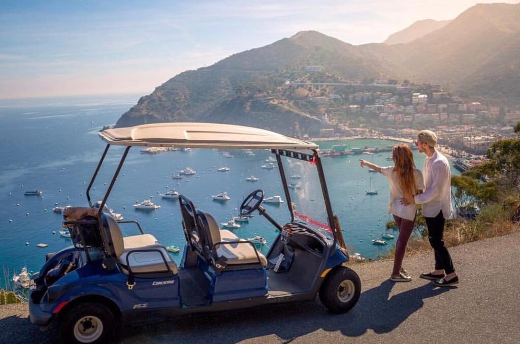 Couple driving a golf cart along coastal road with Avalon Bay view