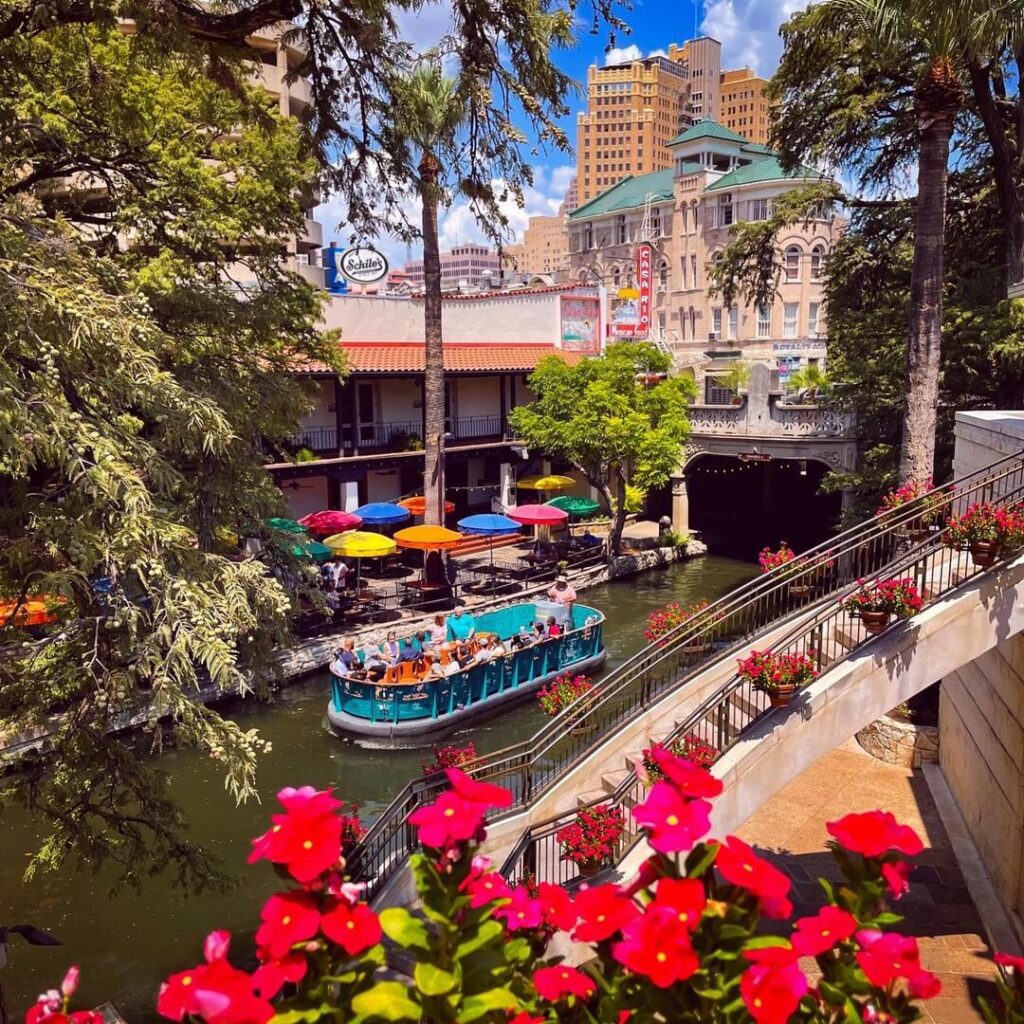 View of San Antonio River Walk at dusk with string lights, café tables, and boats gliding along the canal