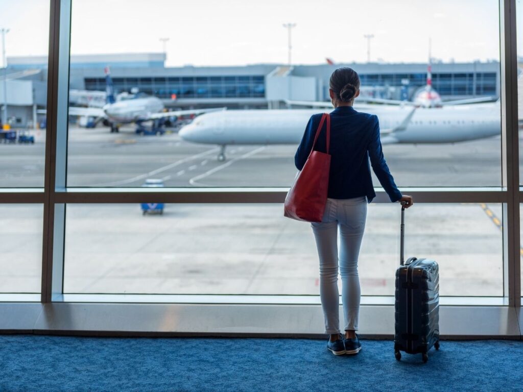 A woman looking out of the airport window with her suitcase beside her	