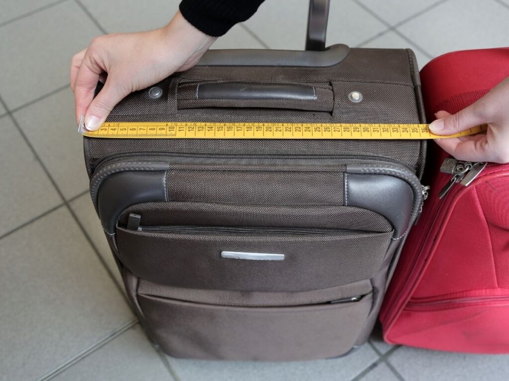 Traveler measuring a carry-on suitcase at home with a tape measure to avoid baggage fees.