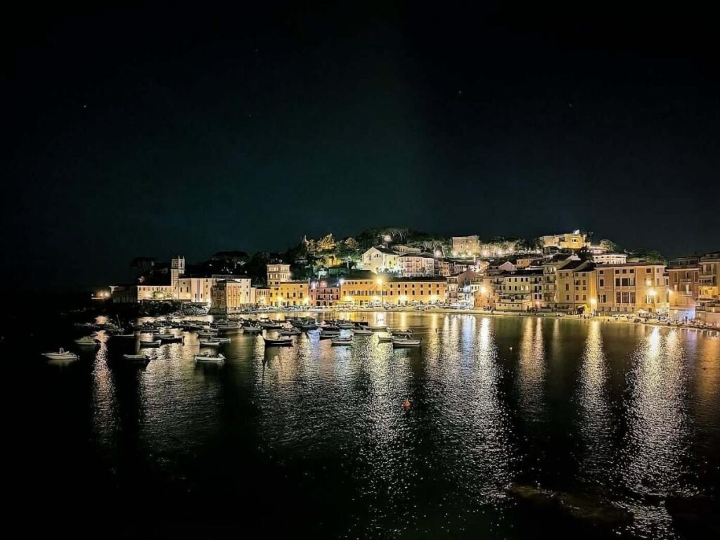Evening scene at Baia del Silenzio with locals strolling the promenade and someone enjoying focaccia on a bench.