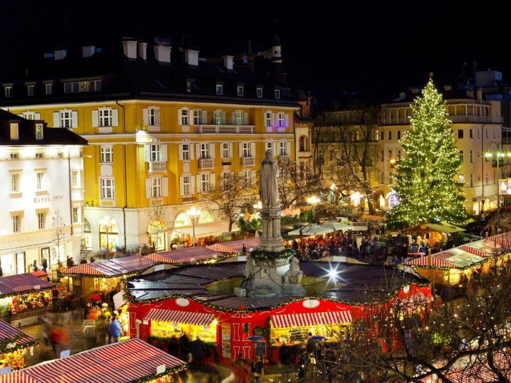 Bolzano street scene with arcaded shops, outdoor market stalls, and mountains in the distance.