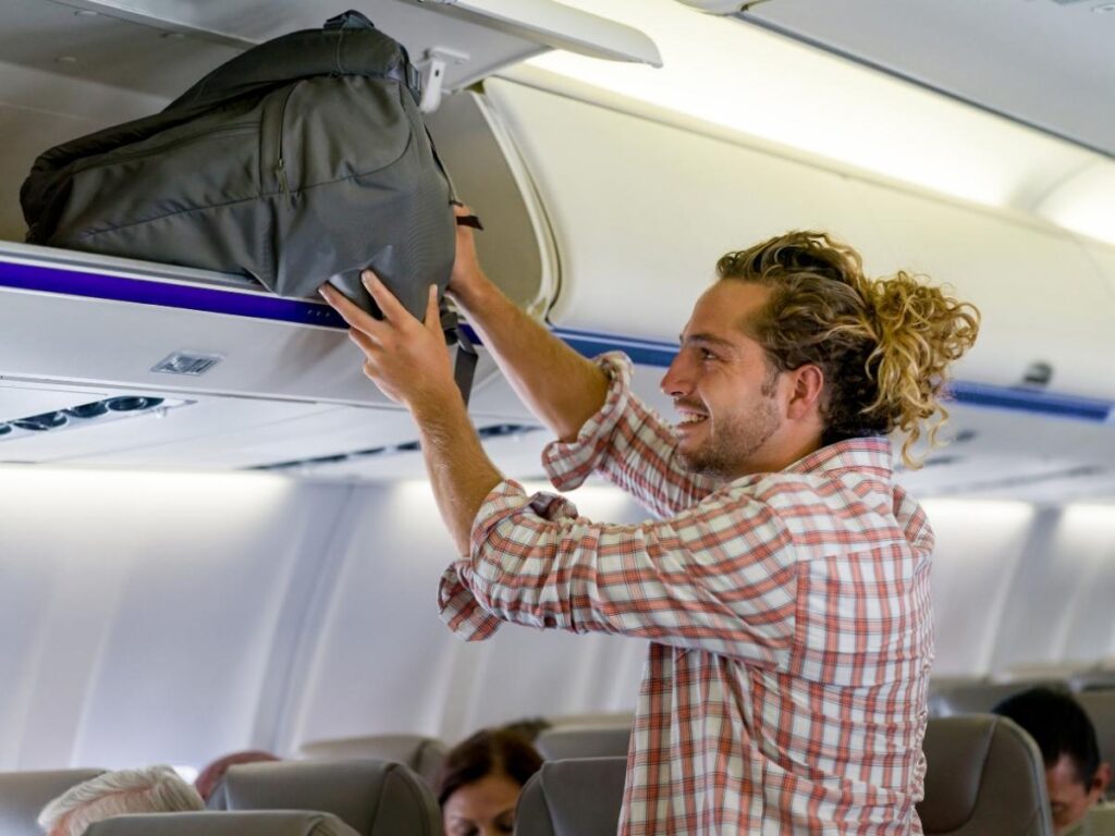 Passenger placing a carry-on bag in the overhead bin to keep legroom free under the seat.