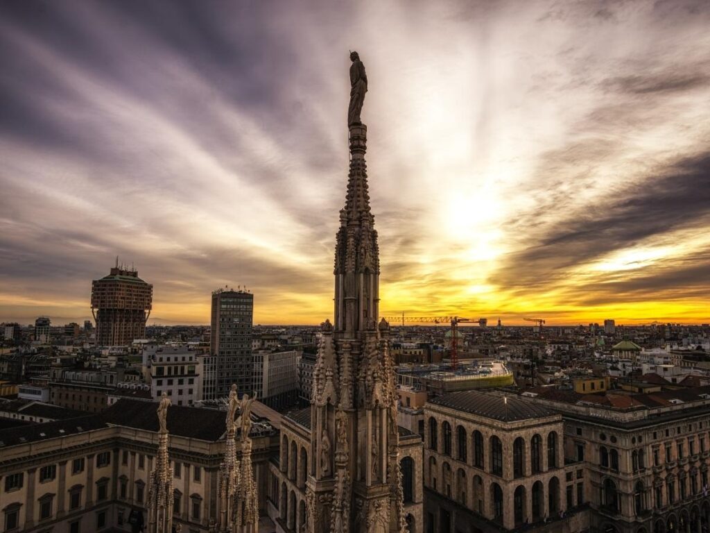 Vertical image of the Duomo rooftop at sunrise with a visitor looking at Milan below — text-safe area for overlay.
