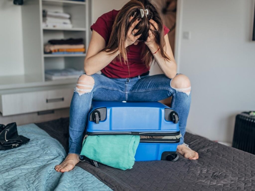 Woman sitting on suitcase trying to close it after packing too many unnecessary items