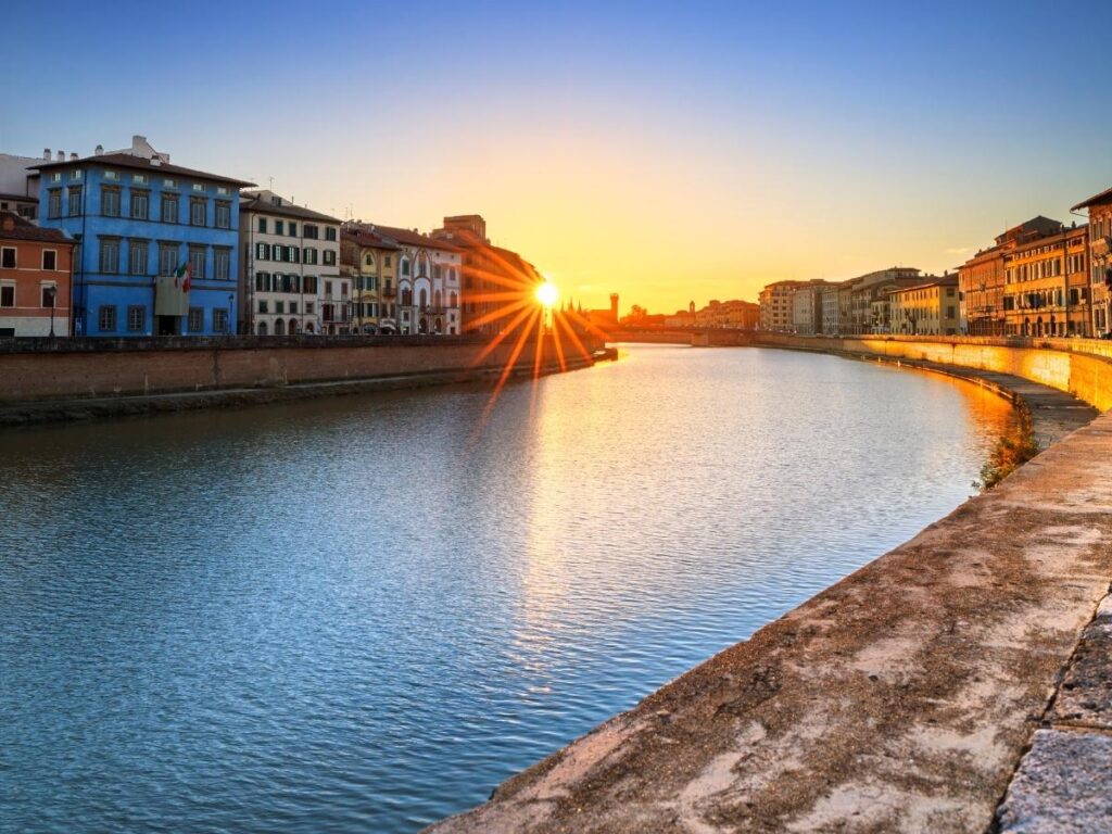Sunset view of Pisa’s Arno River with pastel-colored buildings reflecting in the water.