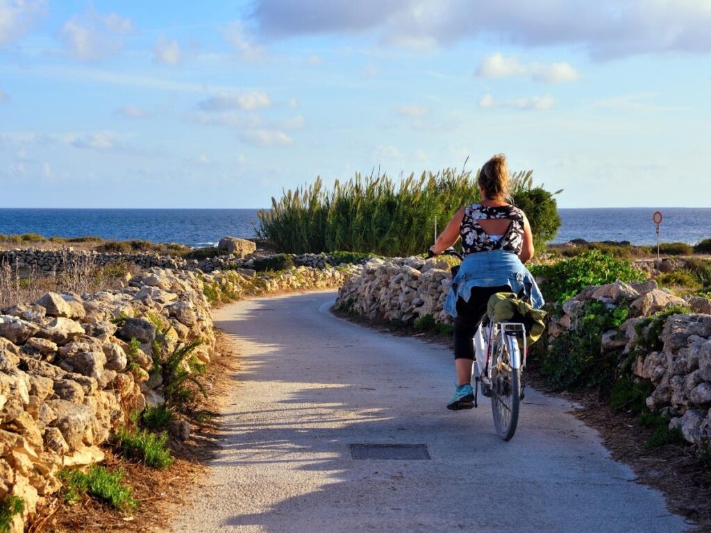 Cyclist on Favignana island with a view of a turquoise bay and rocky coastline.