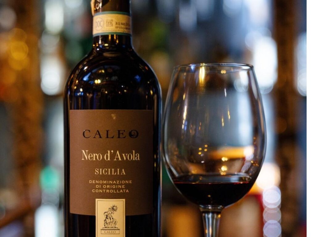 Glass of Nero d’Avola red wine on a rustic wooden table with Sicilian countryside in the background.