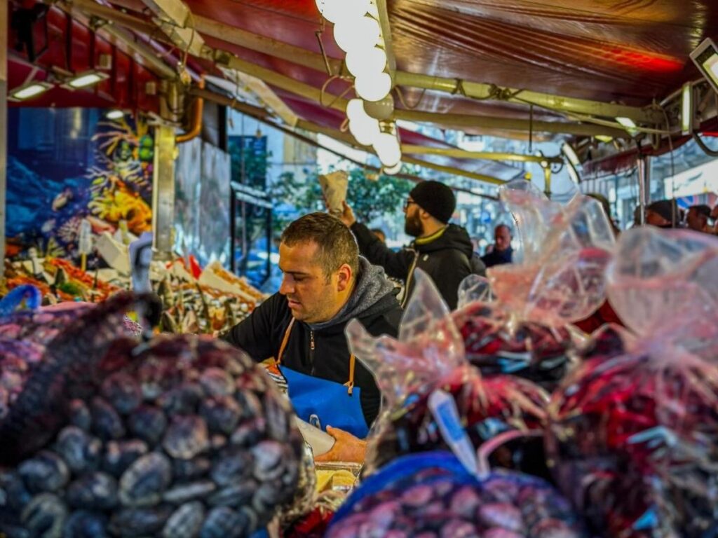 Busy Ballarò market stall in Palermo with vendors, piled citrus and a vendor frying arancini.”