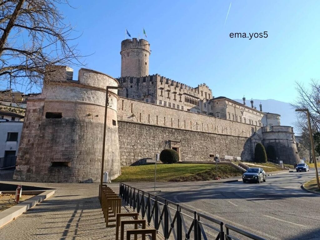 Buonconsiglio Castle rising above Trento’s rooftops, with a blue sky and people in the piazza.