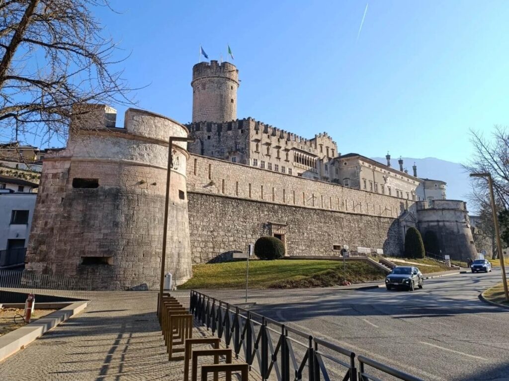 Buonconsiglio Castle rising above Trento’s rooftops, with a blue sky and people in the piazza.