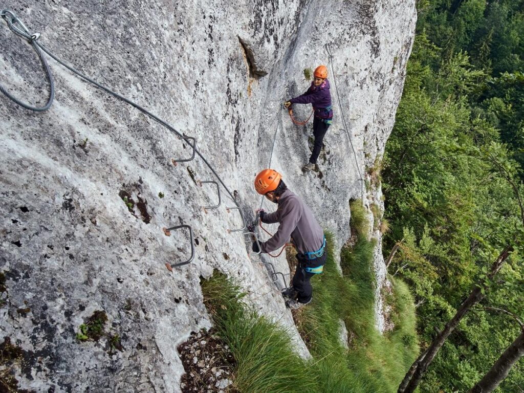 Climber clipped into a via ferrata steel cable on a cliff face with valley views below.