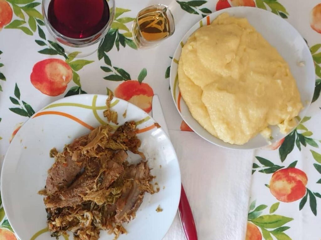 Rustic osteria table with steaming plates of polenta, bread, and homemade grappa served in small glasses.