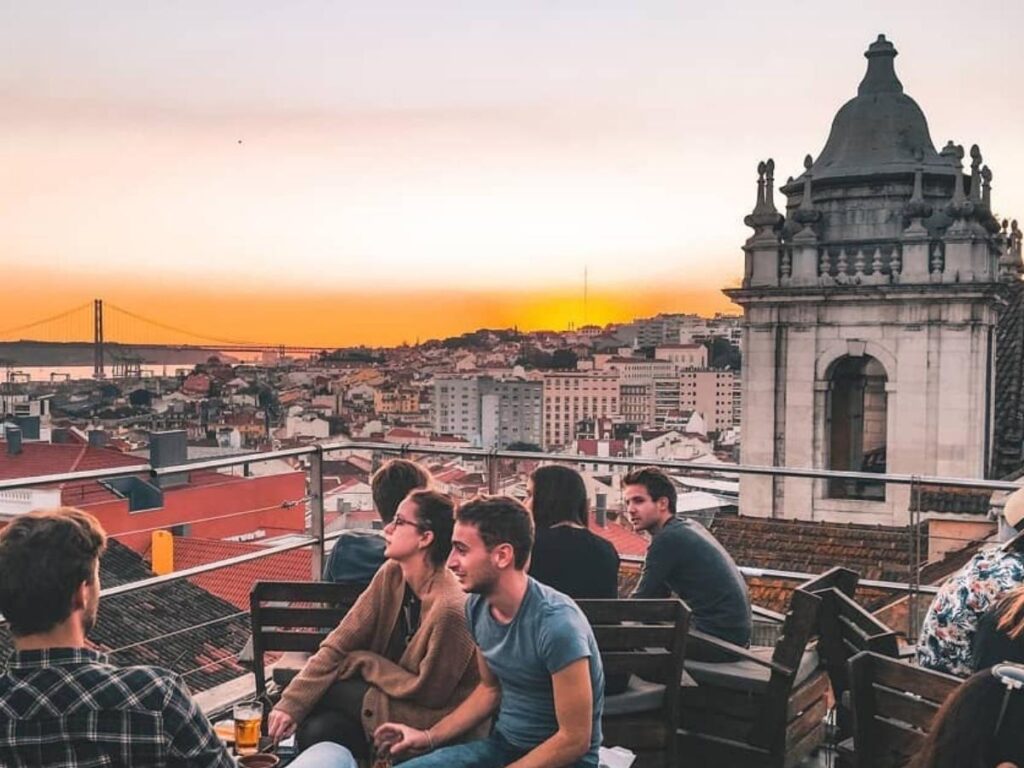 Rooftop bar view over Bairro Alto at golden hour, with people drinking and the city skyline behind.