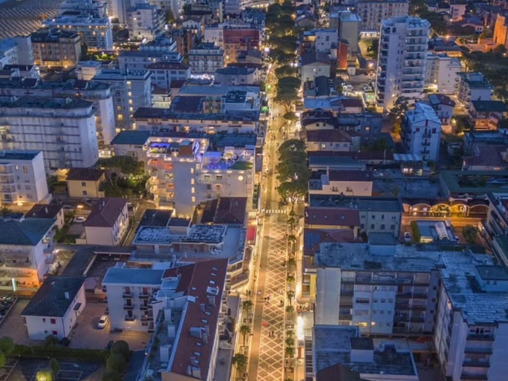 Crowds strolling Via Bafile promenade at dusk with lights and shops.