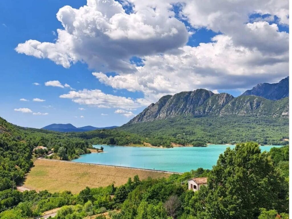 Turquoise waters of Lake Castel San Vincenzo framed by pine trees and distant mountain peaks.