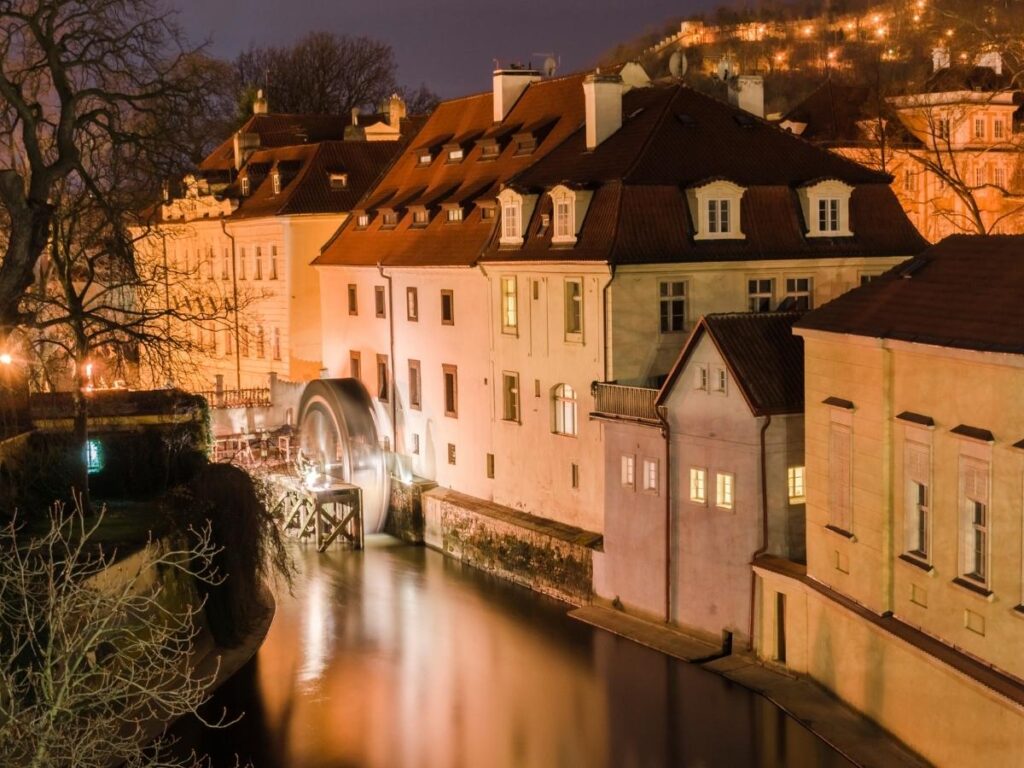 Narrow cobbled lane in Malá Strana leading toward Kampa Island, with pastel facades and a riverside bench