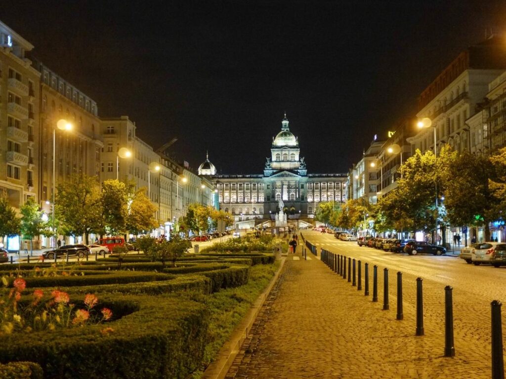 Wenceslas Square at dusk with tram lines, people walking, and city lights coming on in Prague