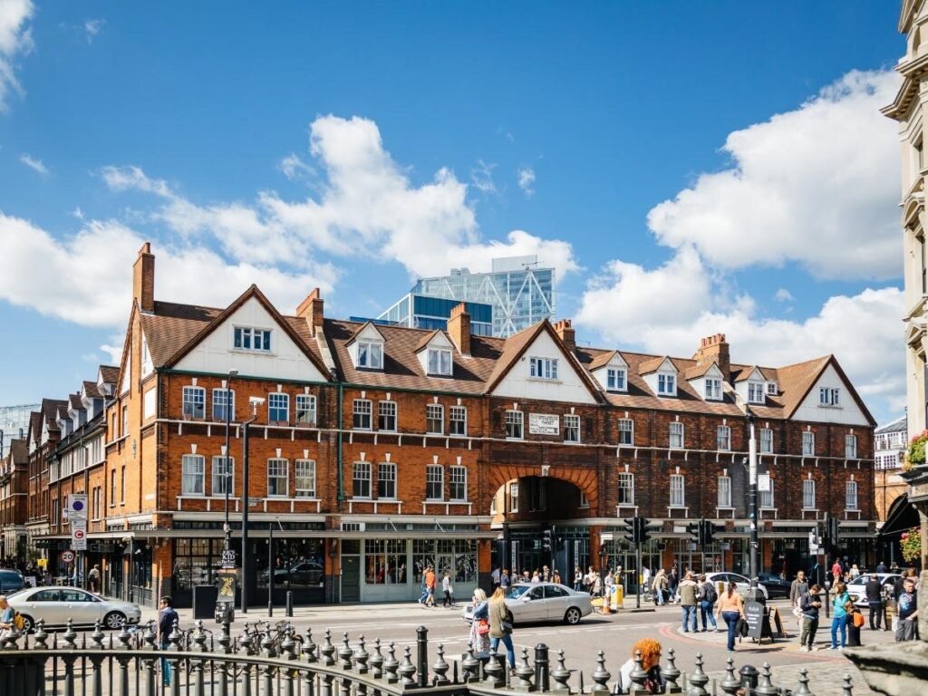 “Entrance of Old Spitalfields Market with shoppers and glass-roofed stalls in East London.”