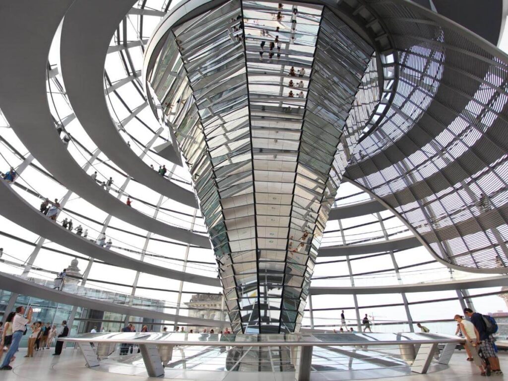 Glass dome of the Reichstag building with visitors walking the spiral ramp and city view in background.
