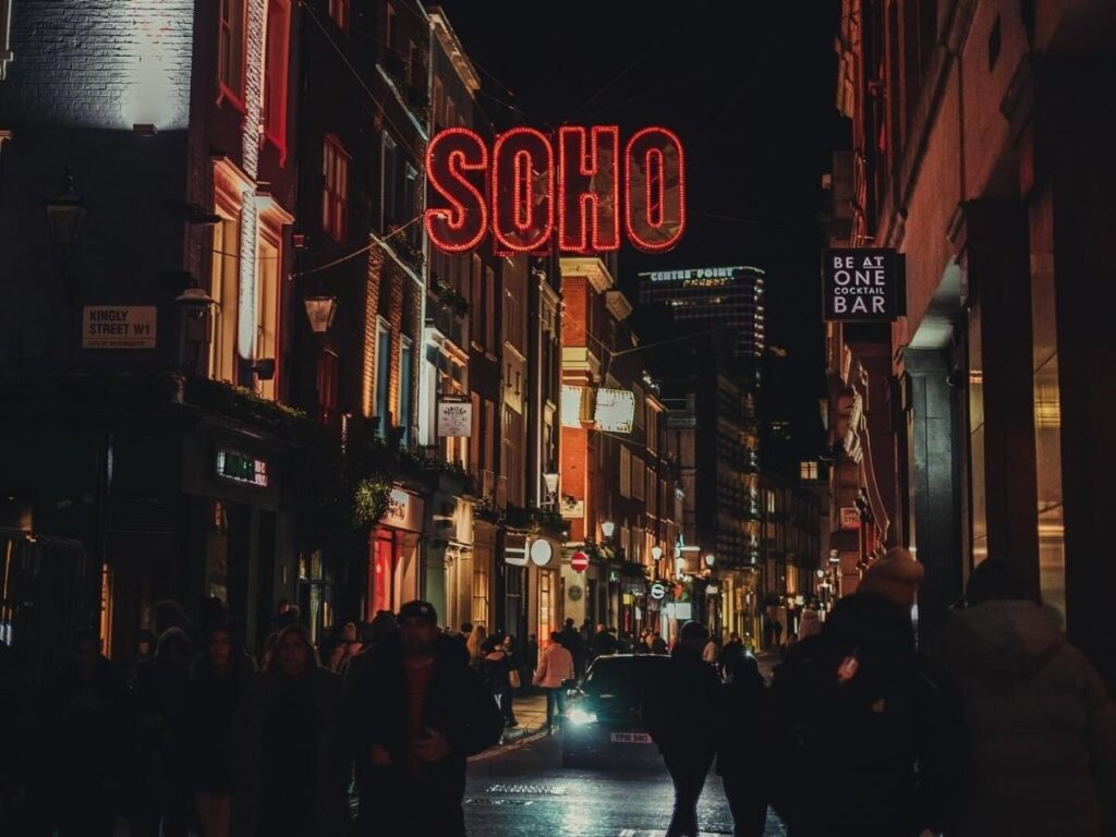 Night scene in Soho, London with glowing theatre signs and people walking along lively streets.