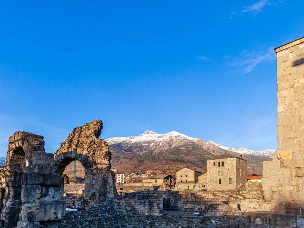 Aosta Roman Theatre outer wall glowing at sunset with the Alps faintly visible behind it.