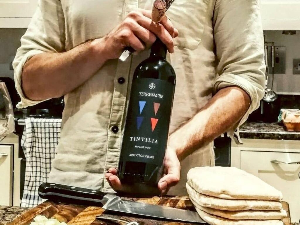 Bottle and glass of Tintilia red wine on a rustic wooden table with vineyards blurred behind.
