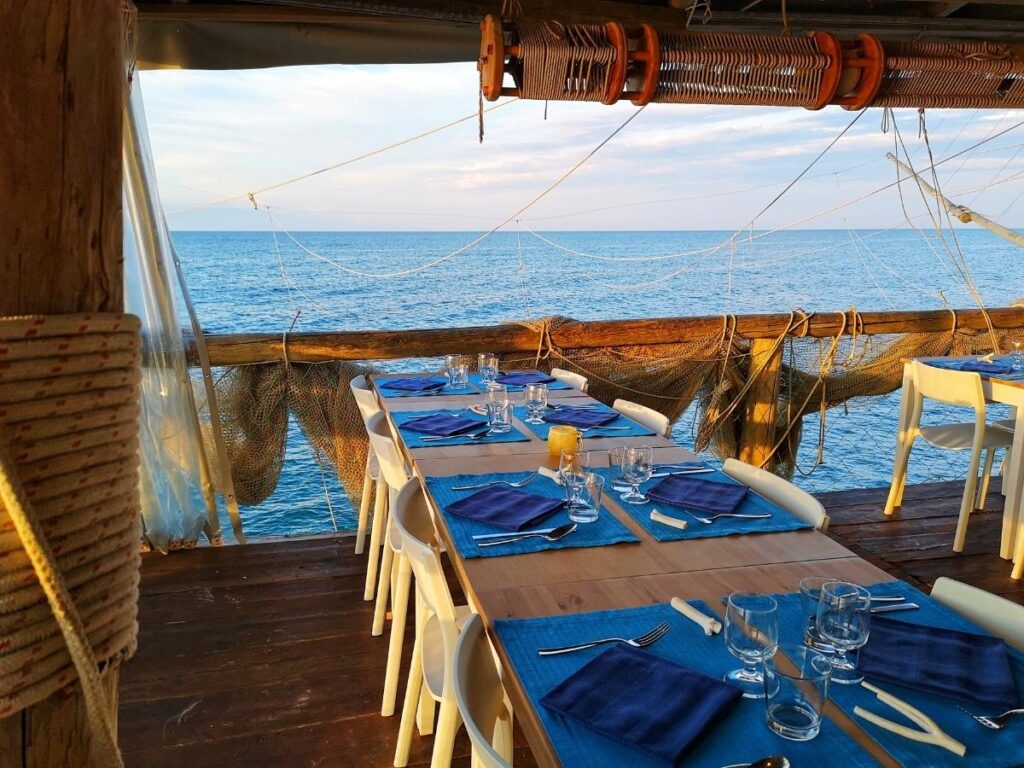 Table set with plates of grilled fish and wine on a trabocco, waves visible below — a seaside meal in Abruzzo