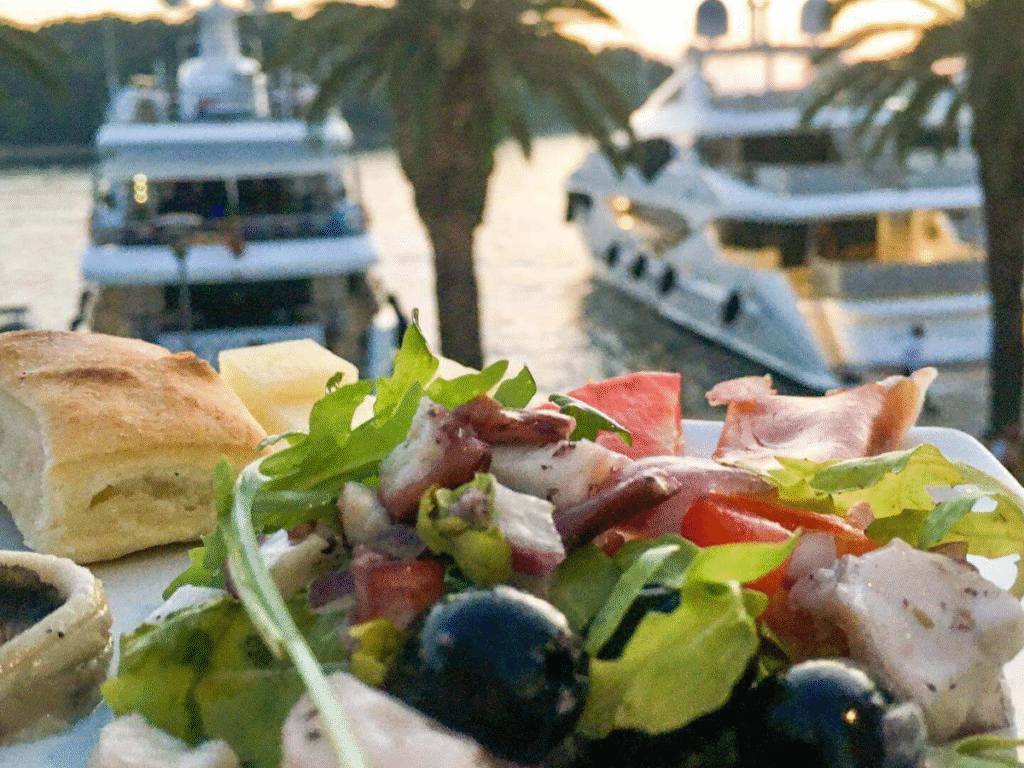 Waterfront table at an outdoor restaurant with a plate of grilled fish, a glass of white wine, and harbour views