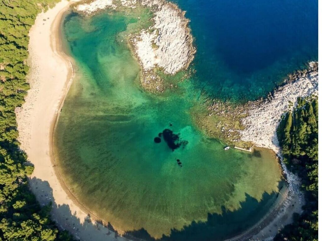 Saplunara sandy bay with pine trees behind the shore and people walking barefoot along the sand