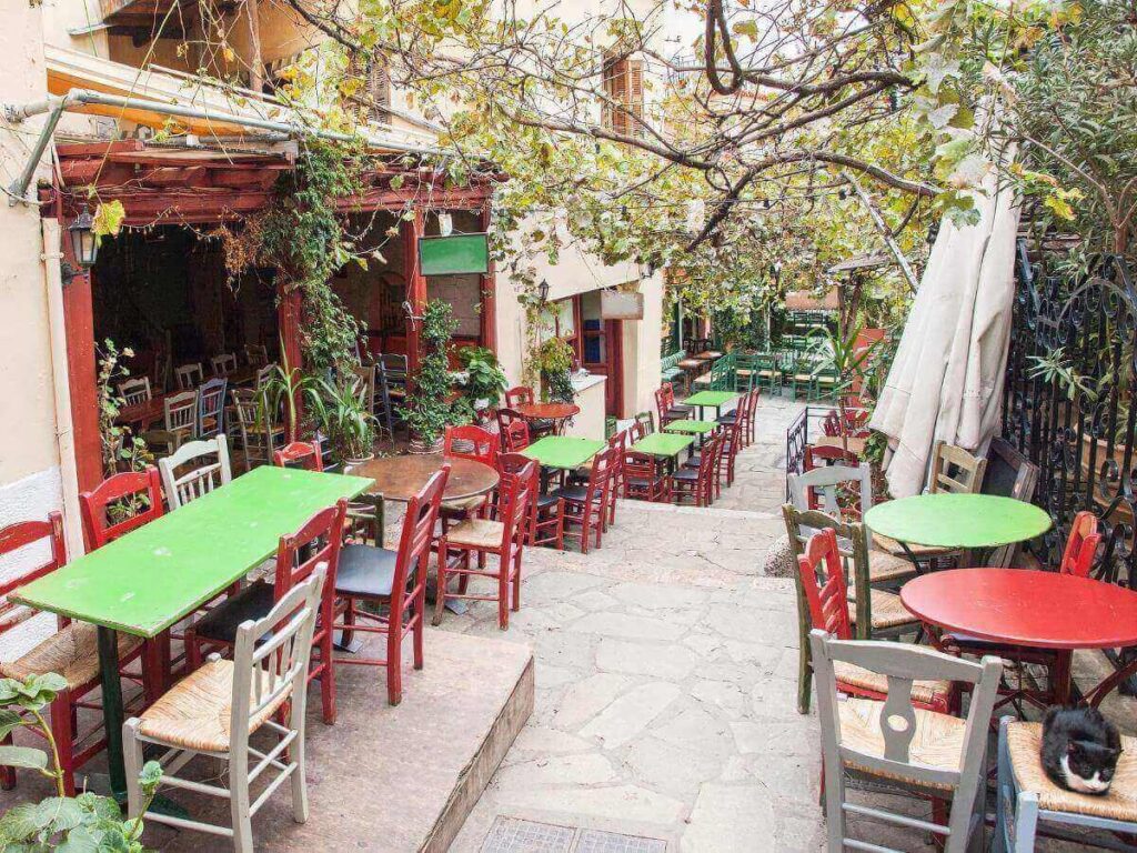 Outdoor cafés and tavernas along a cobbled street in Plaka, Athens, filled with sunlight and flowers.