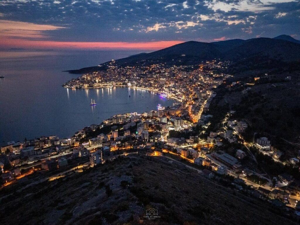 Harbour-side restaurants and tables lit in the evening with fishing boats bobbing in Sarandë”