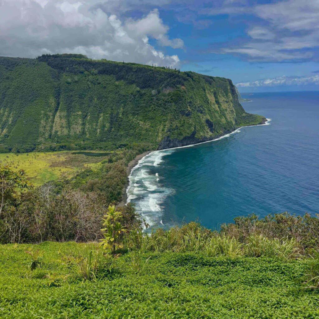 Panoramic view of Waipiʻo Valley with waterfalls, lush cliffs, and a black sand beach