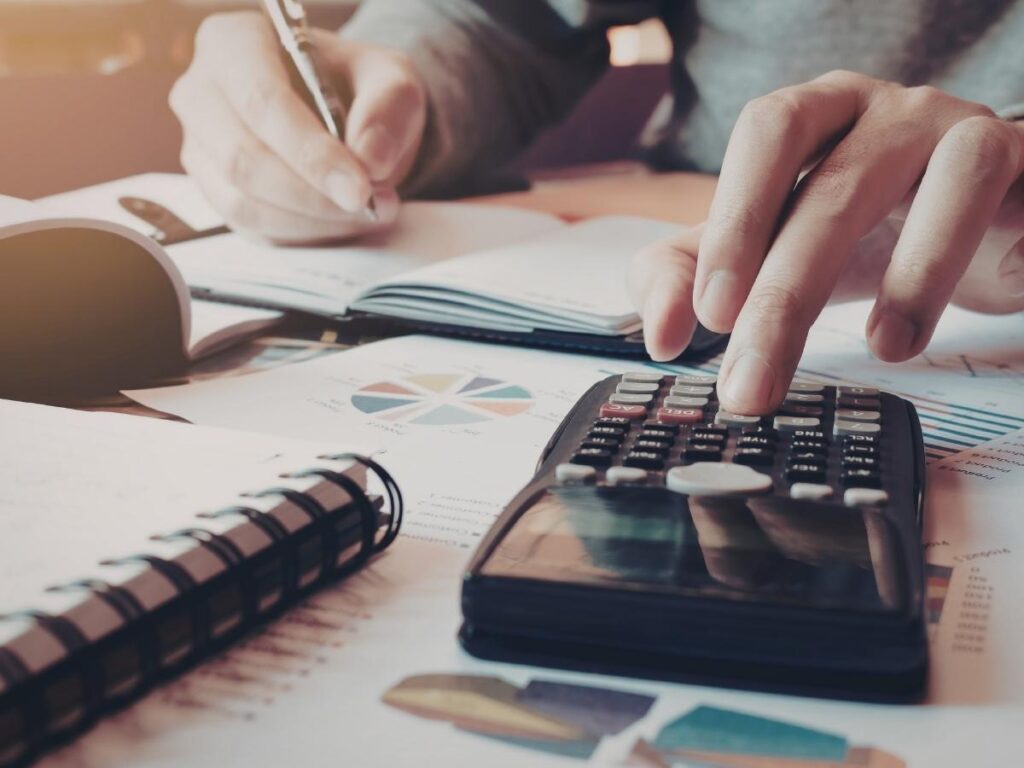 Hands using calculator and writing road trip budget breakdown in notebook