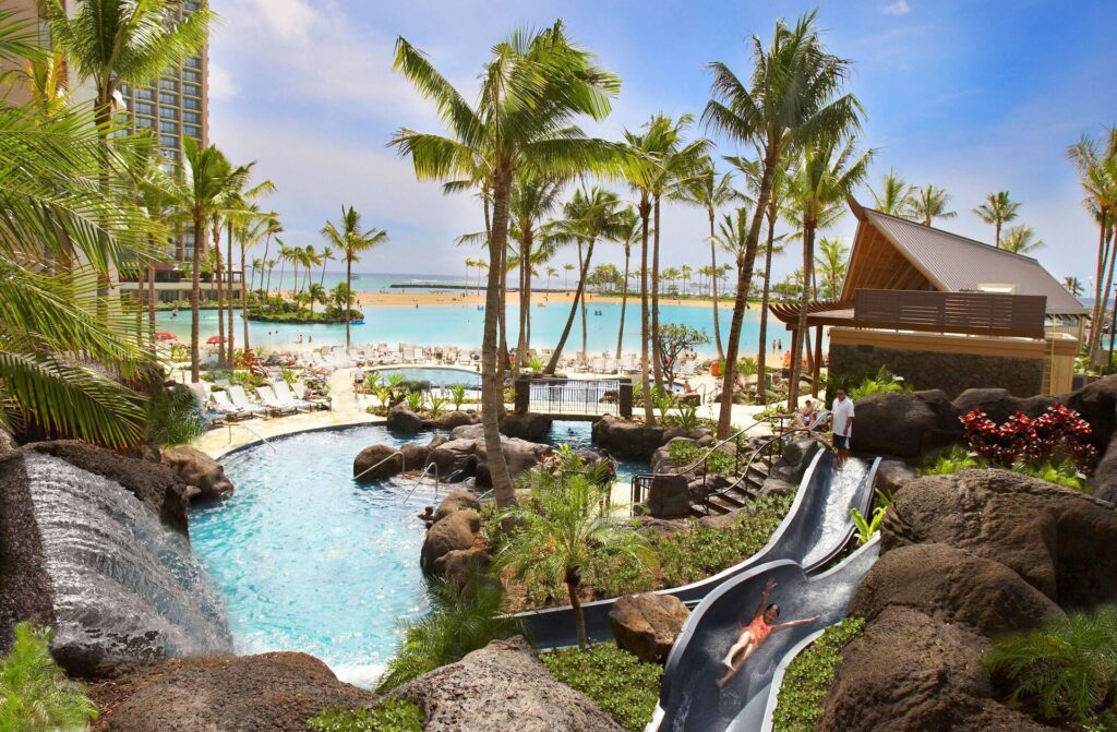 Exterior view of Hilton Hawaiian Village Waikiki Beach Resort in Waikiki, featuring expansive beachfront grounds, multiple resort towers, tropical gardens, pools, and vibrant oceanfront setting.