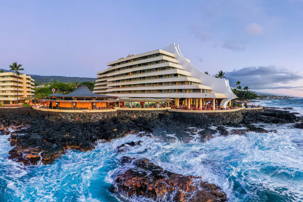 Spectacular oceanfront view of Royal Kona Resort in Kailua-Kona, Hawaii, featuring seaside accommodations, tropical palm trees, lava rock coastline, resort pools, and stunning Pacific Ocean sunsets.