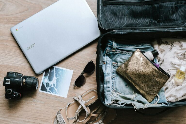 An open suitcase packed with clothes next to a laptop, camera, and sunglasses