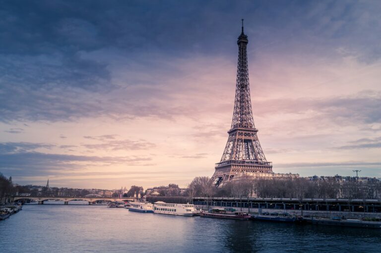 The Eiffel Tower at dusk above the Seine river in Paris, France, with tour boats on the water below.